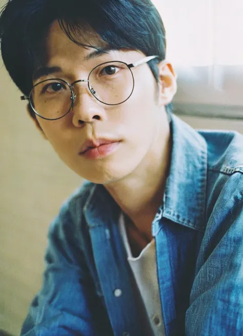 East Asian man, young adult, black hair, round glasses, looking at camera, neutral expression, denim shirt, close-up, plain background, soft lighting, headshot, film grain, soft focus, pensive look, portrait photography, natural skin tone, indoors